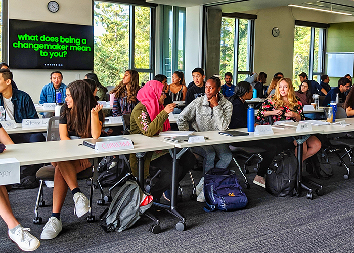 A group of students talking to each other and sitting at a long classroom table