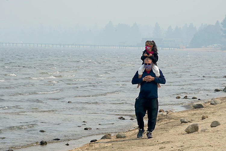A man carries a small child along the beach of Lake Tahoe. They are both wearing face masks, and the air is cloudy with wildfire smoke.