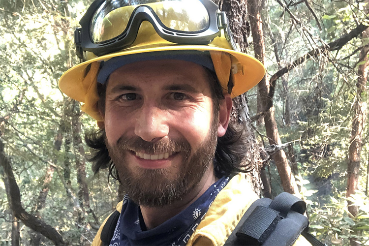 Peter Nelson, an assistant professor at Berkeley, is dressed in firefighter clothing while helping to battle the Walbridge Fire in 2020, in Northern California. The bearded professor has got on a yellow helmet with goggles attached, a navy bandana around his neck, and is carrying gear.