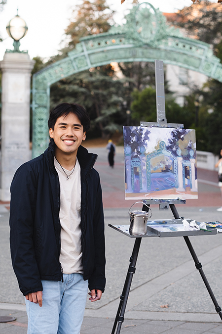 Painter Jake Merioles, wearing a T-shirt, jeans and a fleece jacket, stands next to a painting he's done on Sproul Plaza of Sather Gate.