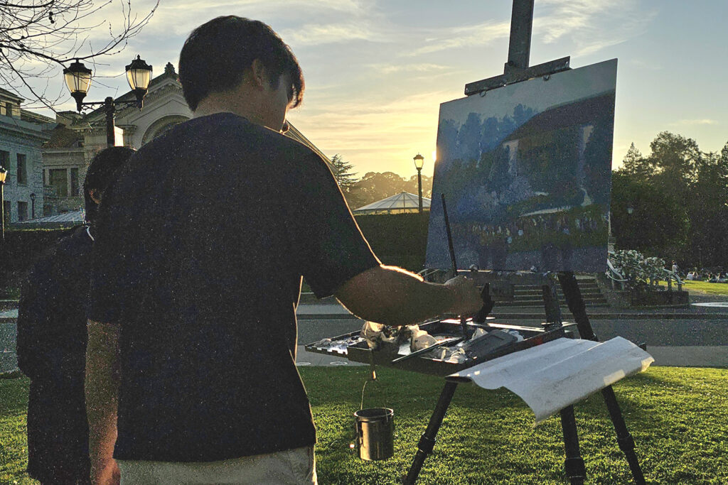 Jake Merioles, a student painter, stands near University Library as the sun begins to set, holding a paintbrush and working on a painting sitting horizontally on an easel in front of him.