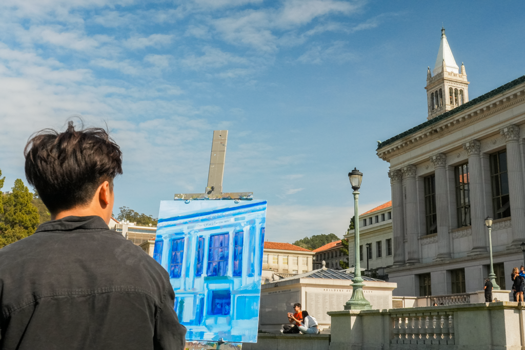 Jake Merioles, a student painter, stands with his back to the camera at an easel in Memorial Glade, working on a painting of University Library. The Campanile is sticking up over the top of the library and the sky is blue with small white clouds. Merioles is wearing a black shirt.