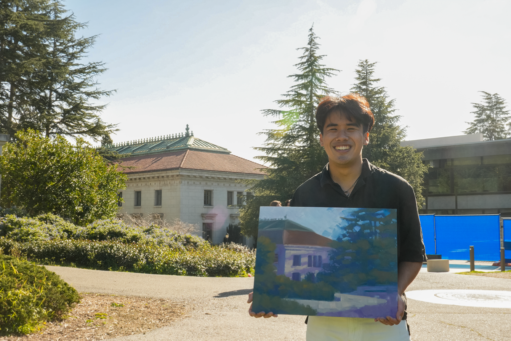 Jake Merioles, a student painter, holds an oil painting of California Hall while he stands a distance from the hall on a campus walkway. He is smiling and wearing off white pants and a black short-sleeved shirt.