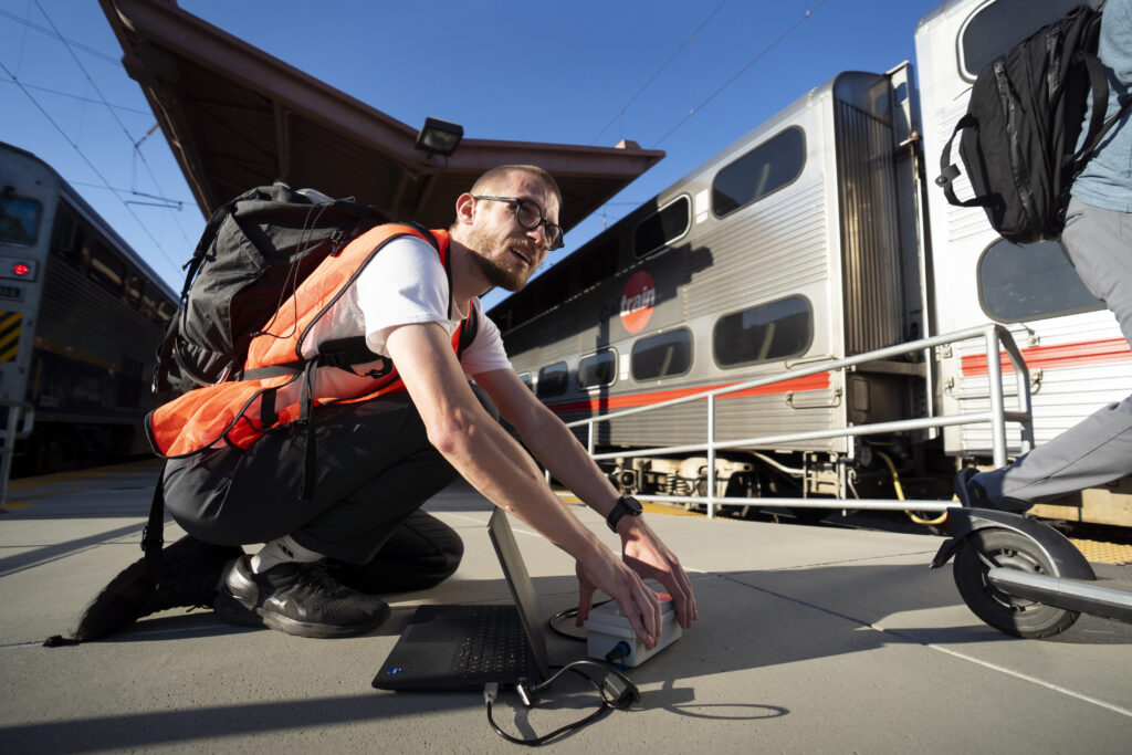 A person kneels on a railway platform in front of a commuter train, plugging a small grey device into a black laptop.