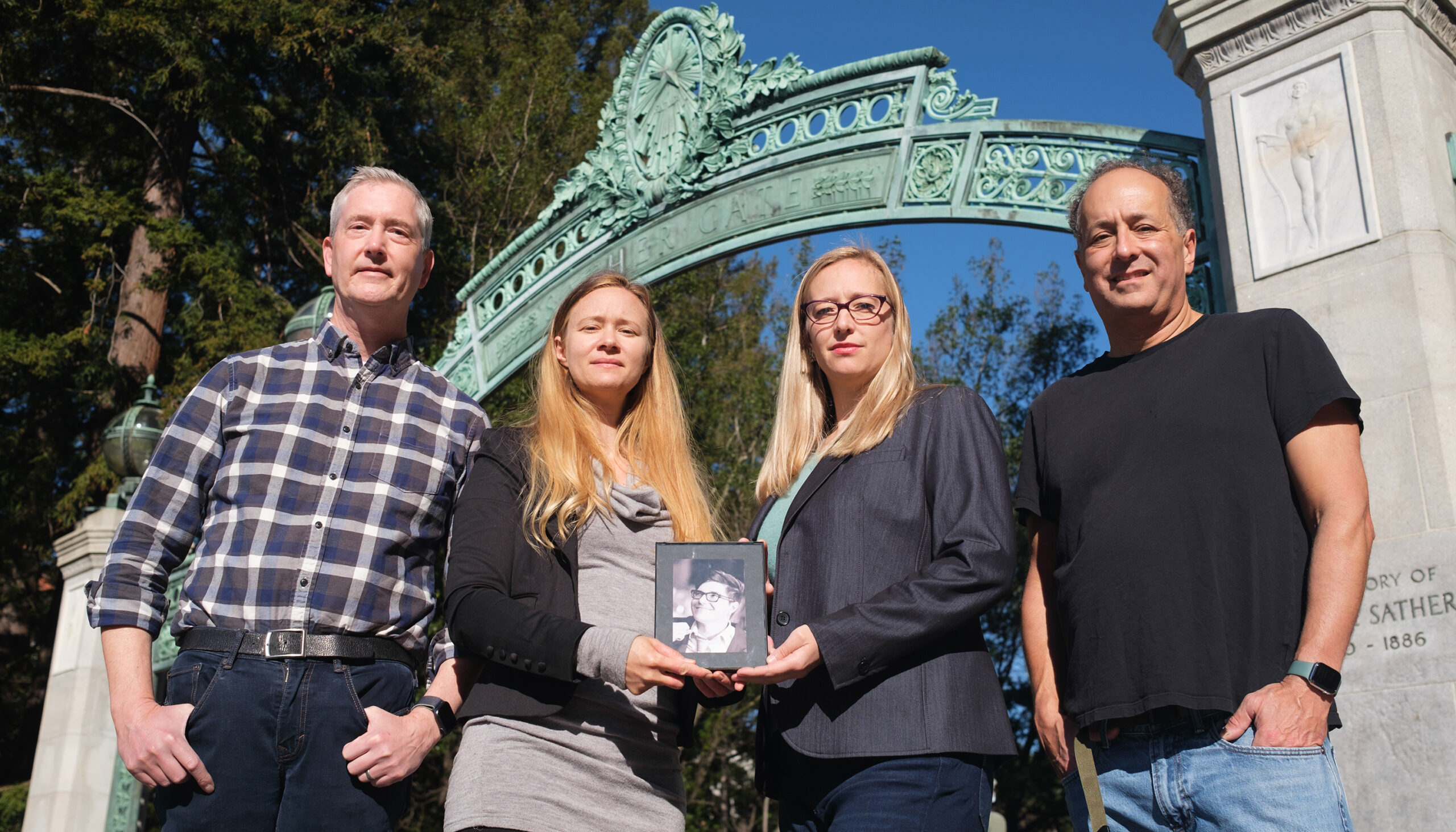 two women and two men stand in front of a green gate holding the photo of a person