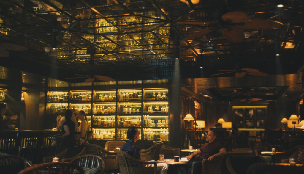 two characters from the show The White Lotus sit at a table in a bar and talk